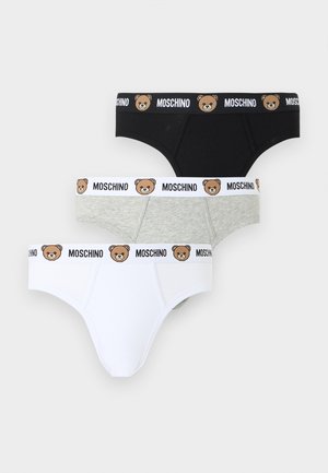 Lihaseline mees seisab paljajalu, seljas hallid aluspüksid valge vööga, millel on karutondijalade näod ja "Moschino" logo, neutraalne taust.