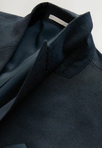 Námornícky blazer s textúrovaným povrchom, s reverzovým golierom a hladkým interiérom. Viditeľné detaily šitia pozdĺž okrajov.