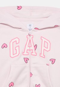 Ανοιχτό ροζ φούτερ με φερμουάρ, με την επιγραφή "GAP" σε μεγάλα ροζ γράμματα, διακοσμημένο με μικρά ροζ μοτίβα καρδιάς και με μπροστινή τσέπη. Μαλακή υφή.