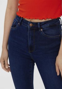 Jeans de tiro alto de color azul oscuro con una textura suave, que presentan un cierre frontal con botón y detalles de costura en naranja.