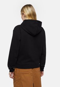 Felpa nera con cappuccio regolabile, polsini e orlo a costine, realizzata in tessuto morbido, indossata sopra una gonna color sabbia con tasche laterali.