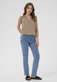 Haut beige à manches courtes avec des épaules froncées, associé à un jean droit bleu clair et des sandales à glisser noires. Design simple et décontracté.