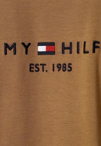 Brun bomuldsstof med broderet tekst "MY HILF" og et rektangulært logo i rød, hvid og sort over "EST. 1985."