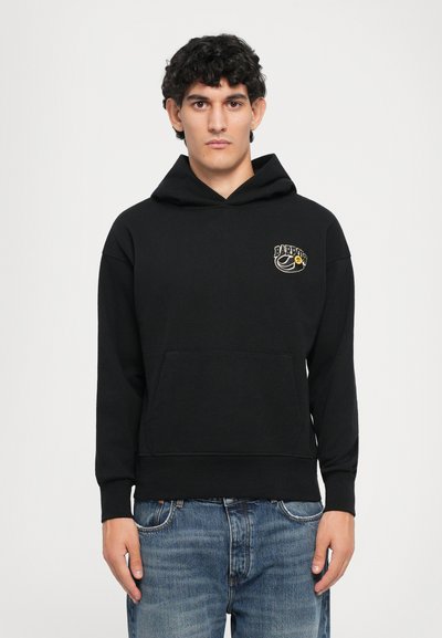 Giovane uomo con capelli ricci che indossa una felpa nera con un piccolo logo sul lato sinistro del petto e jeans blu, in piedi contro uno sfondo semplice.