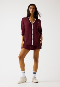 Cardigan bordeaux con scollo a V, dettagli a righe bianche e blu navy, due tasche frontali, abbinato a una gonna plissè e sneakers bianche.