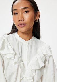 Blusa bianca plissettata con strisce verticali, colletto alto e chiusura con bottoni. Tessuto morbido con volant pronunciati sulle spalle.
