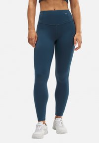 Leggings de talle alto en color teal, elaborados con material elástico, que presentan una textura suave y detalles de costura mínimos. Combinados con zapatillas blancas.