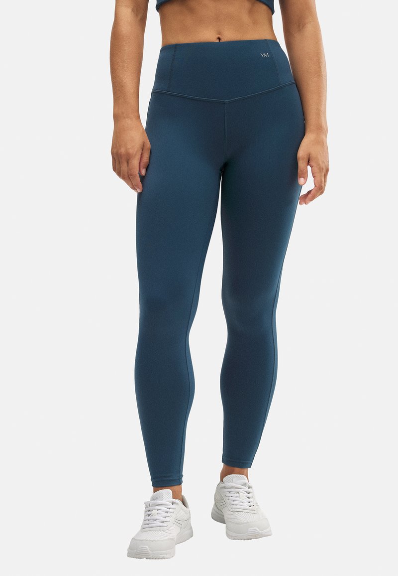 Leggings de talle alto en color teal, elaborados con material elástico, que presentan una textura suave y detalles de costura mínimos. Combinados con zapatillas blancas.