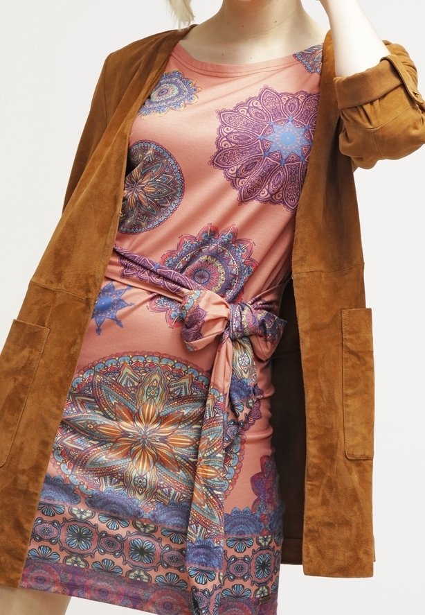Femme portant une robe rose avec des motifs mandala multicolores et une taille nouée, superposée d'une veste en daim marron avec de grandes poches avant.