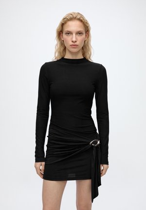 Jeune femme aux cheveux blonds portant une mini-robe noire moulante à manches longues avec un détail froncé et une boucle argentée sur la hanche gauche.