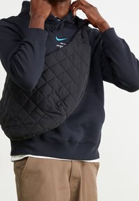 Svart kviltad axelväska med rundad form, med mjukt material, bärs över en marinblå huva med en liten logotyp, kompletterad av tanfärgade byxor.