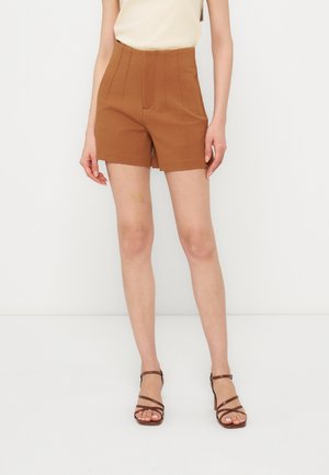 Kvinde iført brune shorts med høj talje kombineret med beige top og brune strop-sandaler med hæl, stående mod en hvid baggrund.