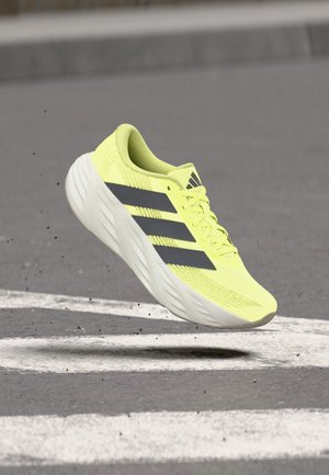 Scarpa da corsa gialla brillante con strisce nere che si libra sopra l'asfalto con segnaletica stradale bianca.