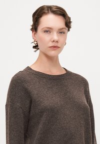 Brun strikkegenser med rund hals og lett løs passform. Myk tekstur og synlig strikkemønster. Modellen har på seg gull hoops.