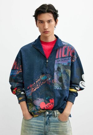 Junger Mann trägt ein blaues Sweatshirt mit halbem Reißverschluss, bunten Mickey-Mouse-Grafiken und ausgewaschenen Jeans, Hände in den Taschen, vor weißem Hintergrund.