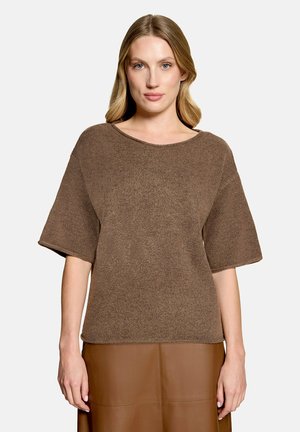 Femme aux cheveux blonds portant un haut en maille marron à manches courtes et une jupe en cuir marron, debout sur un fond clair uni.