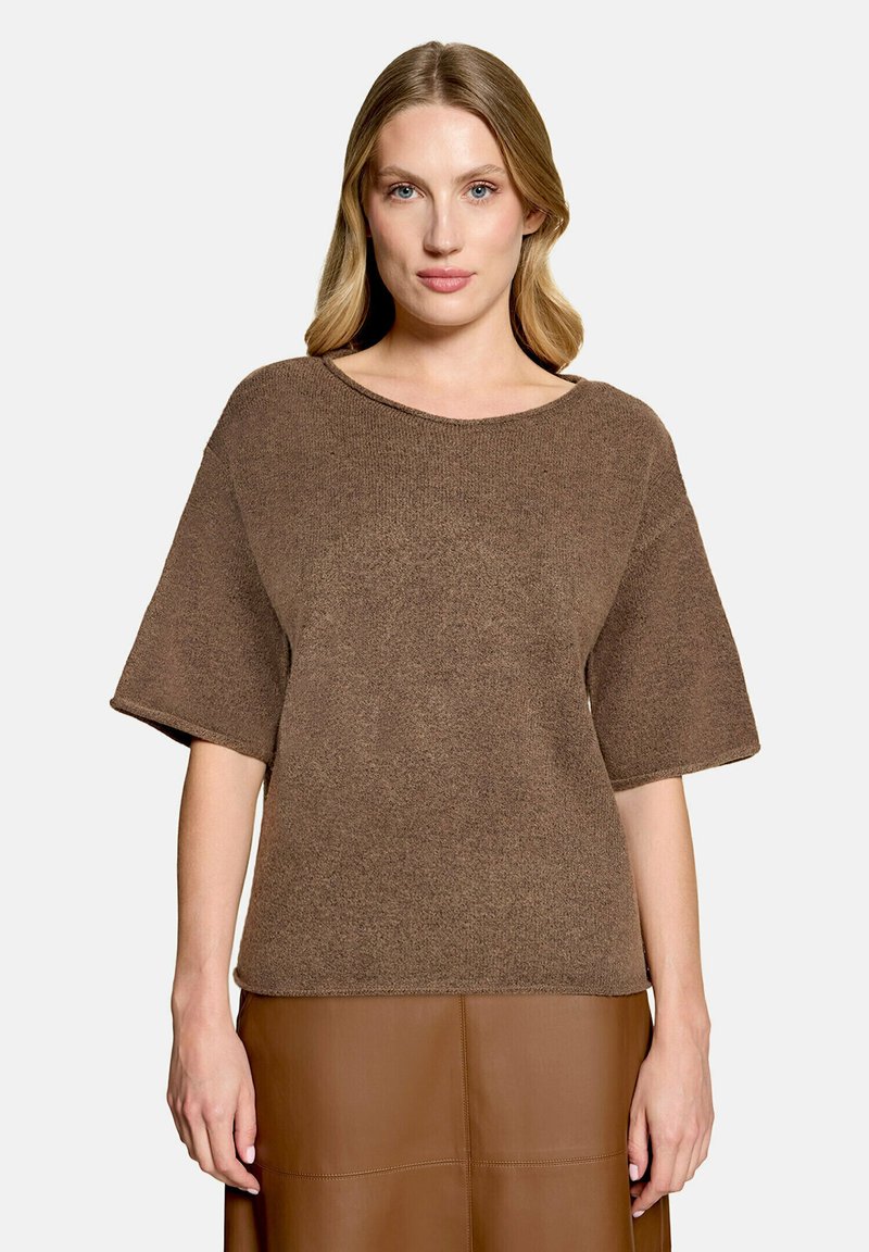 Femme aux cheveux blonds portant un haut en maille marron à manches courtes et une jupe en cuir marron, debout sur un fond clair uni.