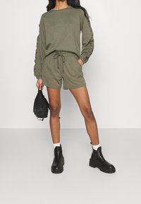 Felpa verde oliva con maniche arricciate, abbinata a pantaloni corti con coulisse dello stesso colore. Il modello indossa stivali neri alla caviglia e tiene in mano una piccola borsa nera.