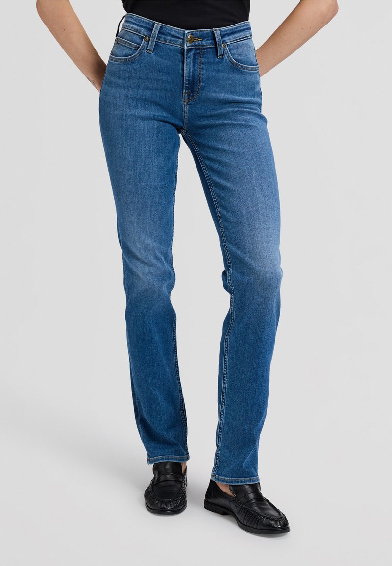 Personne portant un jean bleu droit et des mocassins noirs, debout les mains derrière le dos devant un fond gris uni.