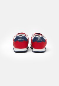 Baskets New Balance rouges avec un accent en mesh bleu au niveau du talon, doublure blanche et semelle en caoutchouc noir. Logo affiché à l'arrière.