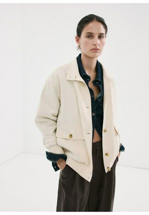 Mujer que lleva una chaqueta color crema sobre una camisa vaquera oscura y pantalones marrones holgados, de pie con las manos en los bolsillos contra un fondo blanco liso.