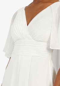 Weißes Kleid mit einem strukturierten, plissierten Oberteil und weiten, durchsichtigen Ärmeln. Der Taillenbund ist glatt und schafft eine fließende Silhouette.