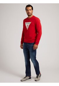 Punainen puuvillapaita, jossa on kolmiomainen "GUESS"-logo, yhdistettynä sinisiin farkkuihin ja harmaisiin lenkkikenkiin, joissa on mustia yksityiskohtia.