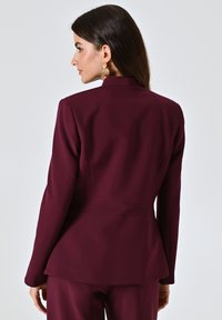 Bordeaux op maat gemaakte blazer met een hoge kraag, gestructureerde pasvorm en een peplumzoom. Glad stof en een minimalistisch ontwerp zonder zichtbare sluitingen.