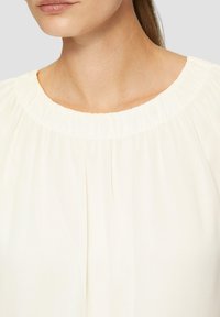 Weiße Bluse mit gerafftem Ausschnitt, weicher Textur und lockerer Passform. Verfügt über einen dezenten Glanz und feine Falten um den Kragen.