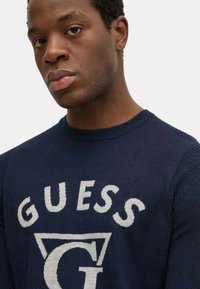 Maglione lavorato a maglia blu navy con un grande logo bianco "GUESS" e un design triangolare. Collo a coste e texture liscia.
