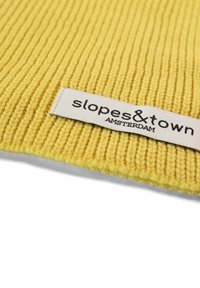Tessuto giallo a righe con un'etichetta bianca che mostra "slopes&town" e "AMSTERDAM" in testo nero. La texture appare morbida e lavorata a maglia.