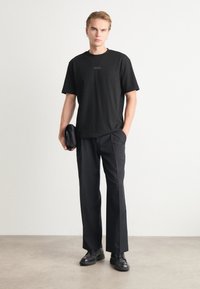 T-shirt en coton noir avec logo gaufré, associée à un pantalon noir ajusté et des chaussures en cuir noires. Le sujet tient un petit sac noir.