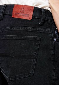 Primer plano del bolsillo trasero de unos vaqueros de mezclilla negros con una etiqueta de marca de cuero marrón "Colours & Sons" cosida en la cinturilla.