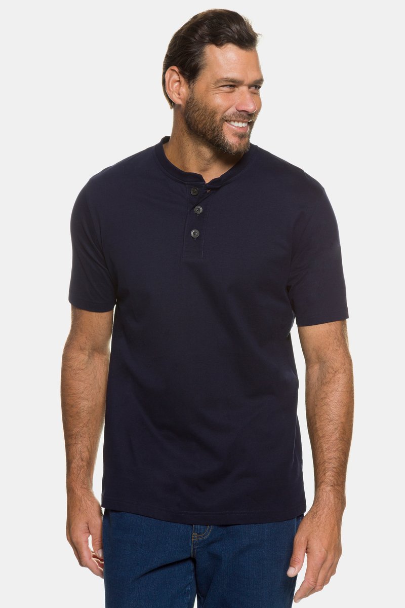 Ein Mann steht und lächelt, trägt ein marineblaues Henley-Shirt mit kurzen Ärmeln und blaue Jeans vor einem schlichten weißen Hintergrund.