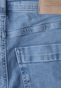 Tissu en denim bleu clair avec des textures variées. Comprend des détails de couture et une petite poche avant. Étiquette en tan texturé à la taille.