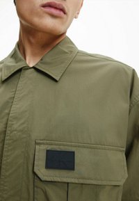 Olivenfarbener Hemdjacke aus glattem Stoff, mit Punktkragen, einer Fronttasche mit schwarzem Calvin Klein Jeans-Logo und sichtbaren Nähten.