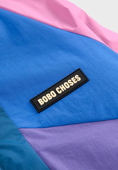 Veste multicolore présentant des panneaux de tissus bleus, roses et violets. Patch logo noir avec texte blanc "BOBO CHOSES" attaché.