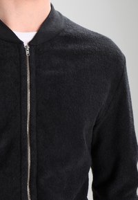 Black bomber jacket made of textured fabric, featuring a zip closure and a ribbed collar. Close-up view highlights material and color.