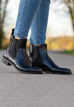 Zwarte gepatineerde leren enkelboots met elastische zijpanelen en decoratieve stiksels, gecombineerd met aangesloten blauwe jeans op een grindpad.