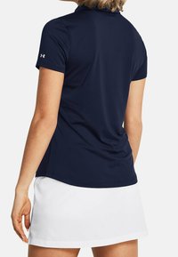 Mörkblå kortärmad polo tröja med en slät textur, som har en krage och subtil logotypdetalj på ärmen. Bärs tillsammans med en vit kjol.