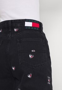 Vaqueros de mezclilla negra con diseños de corazones bordados en rojo, blanco y azul. Incluye una etiqueta "TOMMY JEANS" con un acento de bandera tricolor.
