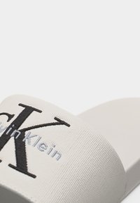 Λευκό σανδάλι slide Calvin Klein με κέντημα λογότυπου σε μαύρο και γκρι σε υφασμάτινο λουράκι, απεικονισμένο σε λευκό φόντο.