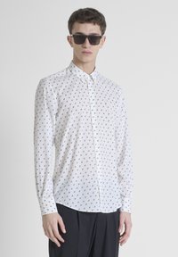 Camicia bianca con piccoli motivi geometrici neri, maniche lunghe e colletto; il modello indossa occhiali da sole scuri. I pantaloni neri completano il look.