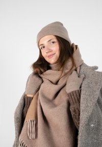 Écharpe et bonnet en mélange de laine beige, gants en tricot texturé, superposés sur un manteau gris. L'écharpe présente des franges. Matériaux doux et chauds.
