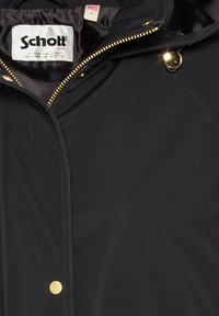 Giacca nera con chiusura a zip e dettagli in oro, dotata di cappuccio e texture morbida. L'etichetta riporta "Schott" con un dettaglio della bandiera USA.