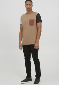 Menns fargeblokkert t-skjorte med en beige kropp, grå og svarte ermer, og en vinrød brystlomme. Kombinert med svarte jeans og svarte sneakers.