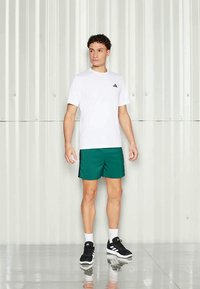 Balta sportinė marškinėliai su mažuoju juodu logotipu krūtinėje, derinami su žaliais šortais. Modelis dėvi juodus sportinius batus ir baltas kulkšnines kojines.