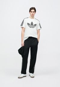 Fehér p�óló fekete mintás Adidas logóval, fekete sportnadrág fehér csíkokkal, fehér tornacipő barna talppal, fekete sapkát tartva.