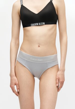 Γυναίκα που φοράει μαύρο Bralette Calvin Klein και γκρι εσώρουχα Calvin Klein στέκεται μπροστά σε απλό φόντο.