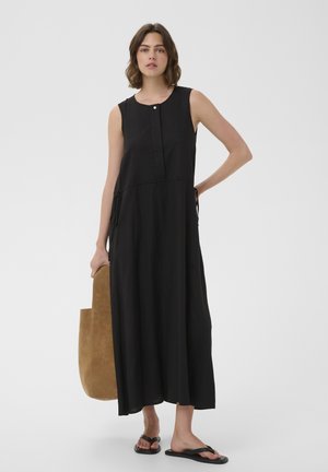 Frau trägt ärmelloses schwarzes Maxikleid mit Seitenschnüren, hält große braune Wildledertasche und trägt schwarze flache Sandalen.